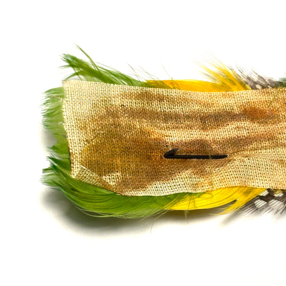 Feathered hair clip with green, yellow, and black feathers on a beige fabric background.