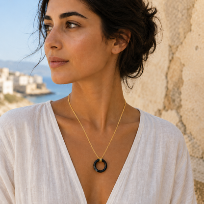 A woman wearing a delicate gold chain necklace with a black circular ring pendant, photographed outdoors against a textured stone wall and sea background.