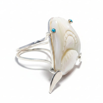 A vintage mother of pearl ring with a shark head design, featuring 18K gold eyes and turquoise fins, on a blue background.